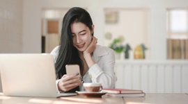  Ein Foto einer fremden Frau, die lächelt, während sie ihr Telefon betrachtet, mit einem Laptop und einer Tasse Kaffee vor ihr